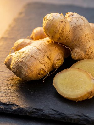 [400×400px | 72dpi | JPG] Macro food photography: organic ginger root with rough golden-tan skin showing fibrous texture, one piece sliced to reveal pale yellow fibrous interior, arranged on dark slate, warm side lighting, editorial macro style, no text