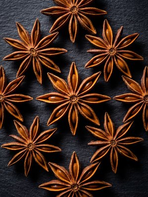 [400×400px | 72dpi | JPG] Macro food photography: perfect whole star anise pods arranged symmetrically on dark slate, showing the eight-pointed star form with seed chambers and woody texture, deep amber-brown color, dramatic overhead lighting, ultra-sharp detail, editorial style, no text